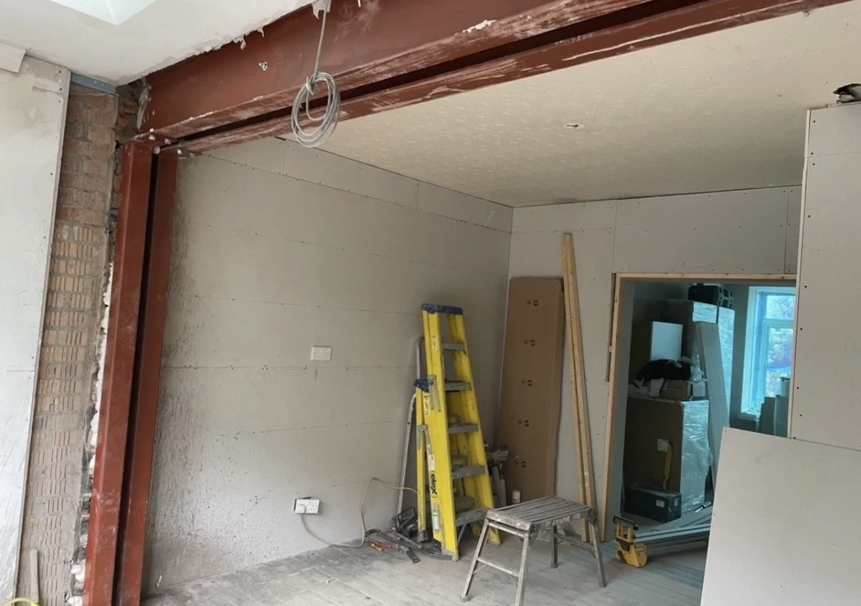 Interior of a room under construction with a steel beam supporting the ceiling, a yellow ladder, wooden planks, and construction tools and materials scattered around.