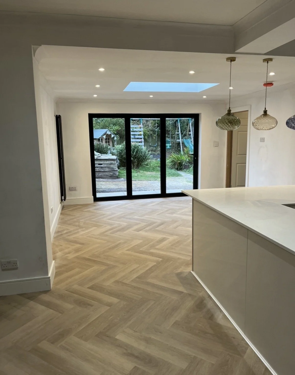 Modern kitchen and dining area with light wooden herringbone patterned flooring, large sliding glass doors opening to a backyard with trees and a playhouse, ceiling recessed lighting, and three hanging pendant lights.