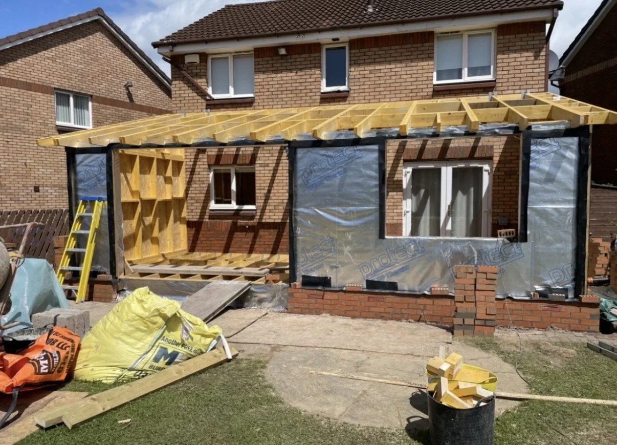 Construction of a small porch or extension on the back of a brick house, with wooden framing and partial wall sheathing, construction materials and tools on the ground.