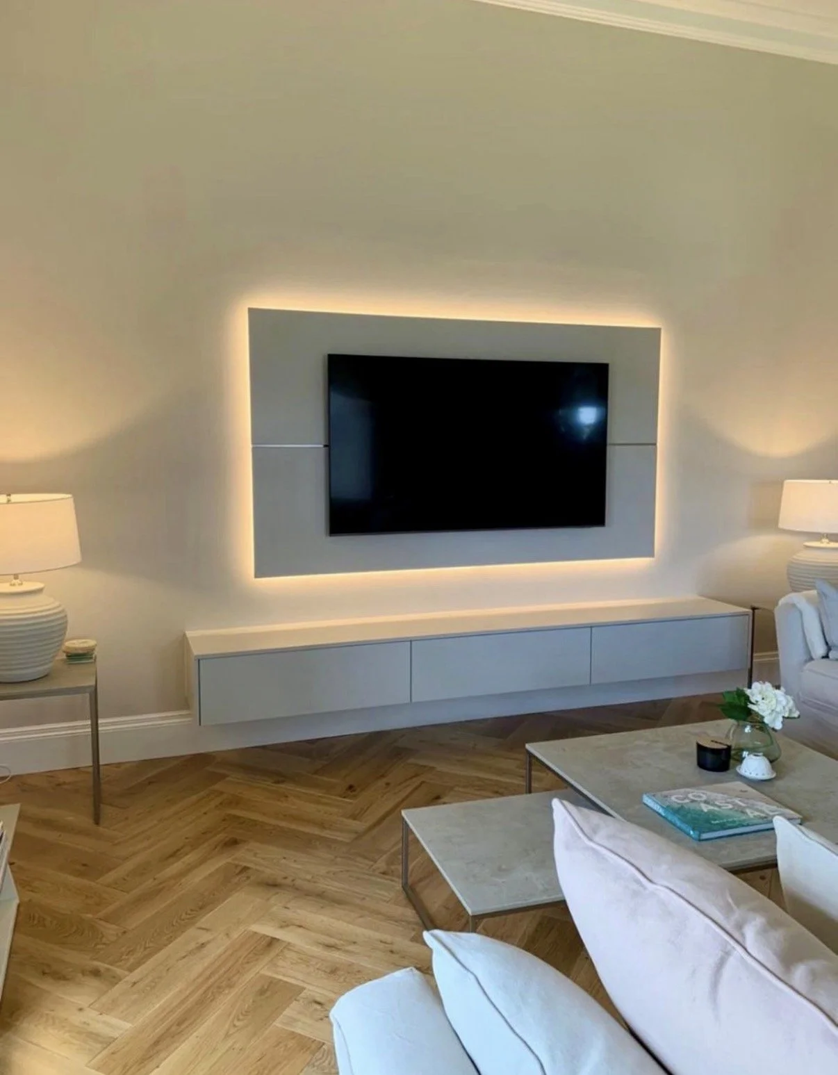 A modern living room with a large flat-screen TV mounted on the wall, surrounded by a backlit panel. There are two white table lamps on side tables, a white console below the TV, a marble coffee table with a flower arrangement, and a white upholstere