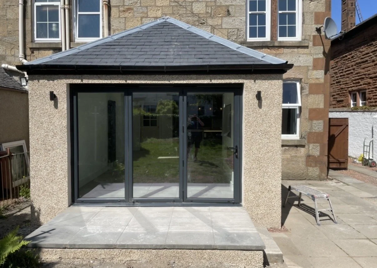 Newly constructed small extension with sliding glass doors and a tiled roof, attached to a stone and brick house, with a small step leading to the patio area.