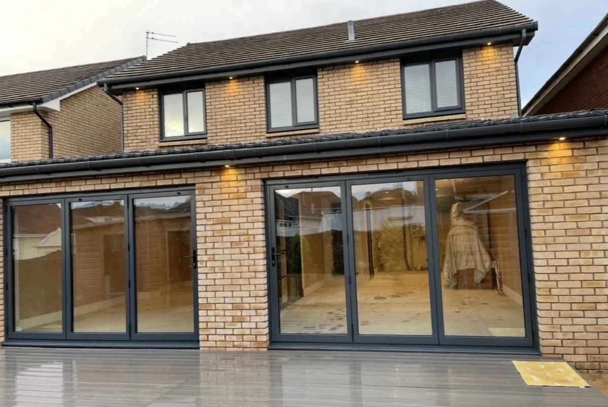 The image shows a modern brick house with large glass sliding doors leading to a wooden deck. The house has a two-story structure with several windows and exterior lights. The interior visible through the doors appears to be unfurnished.