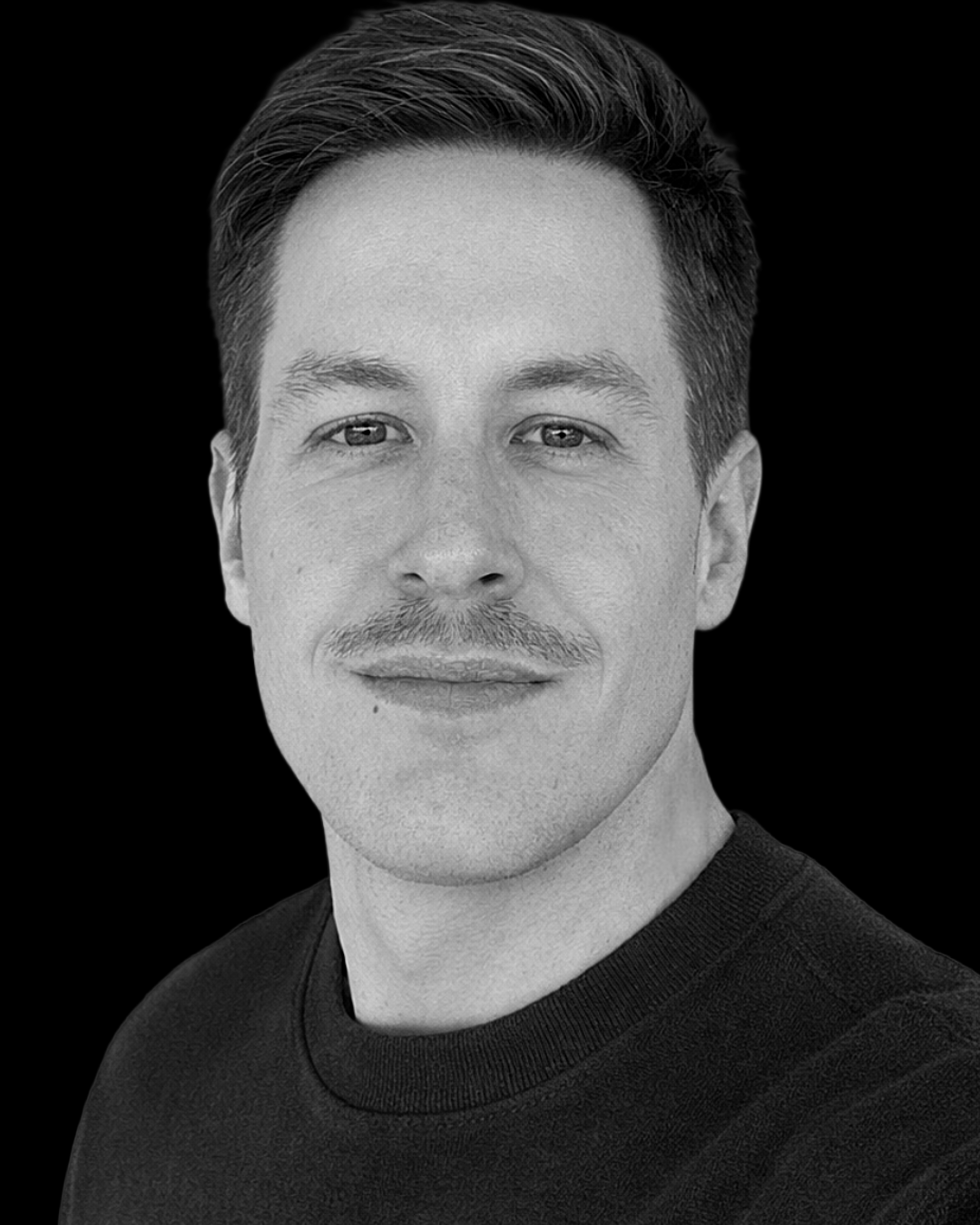 Black and white portrait of a man with short hair, a mustache, wearing a black shirt, looking directly at the camera, against a black background.