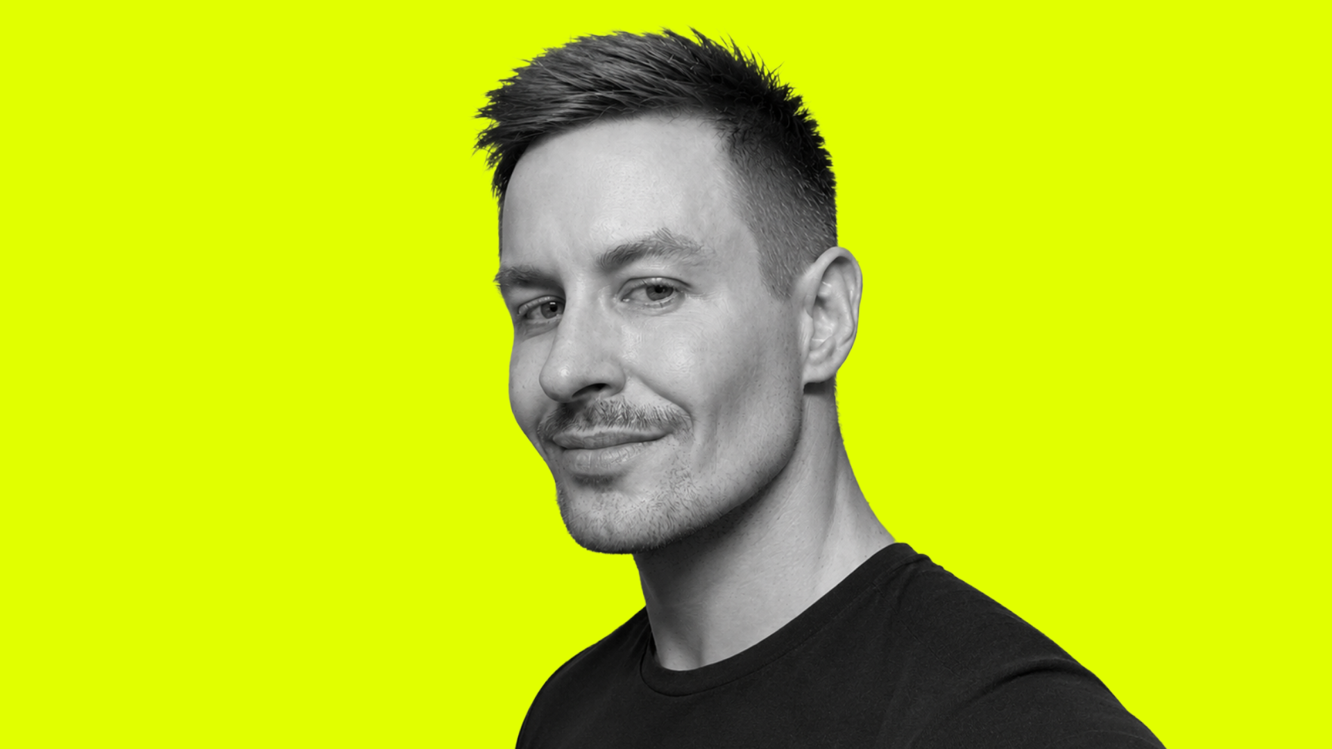Black and white portrait of a young man with short, styled hair, smiling slightly, wearing a black shirt, against a vibrant yellow background.