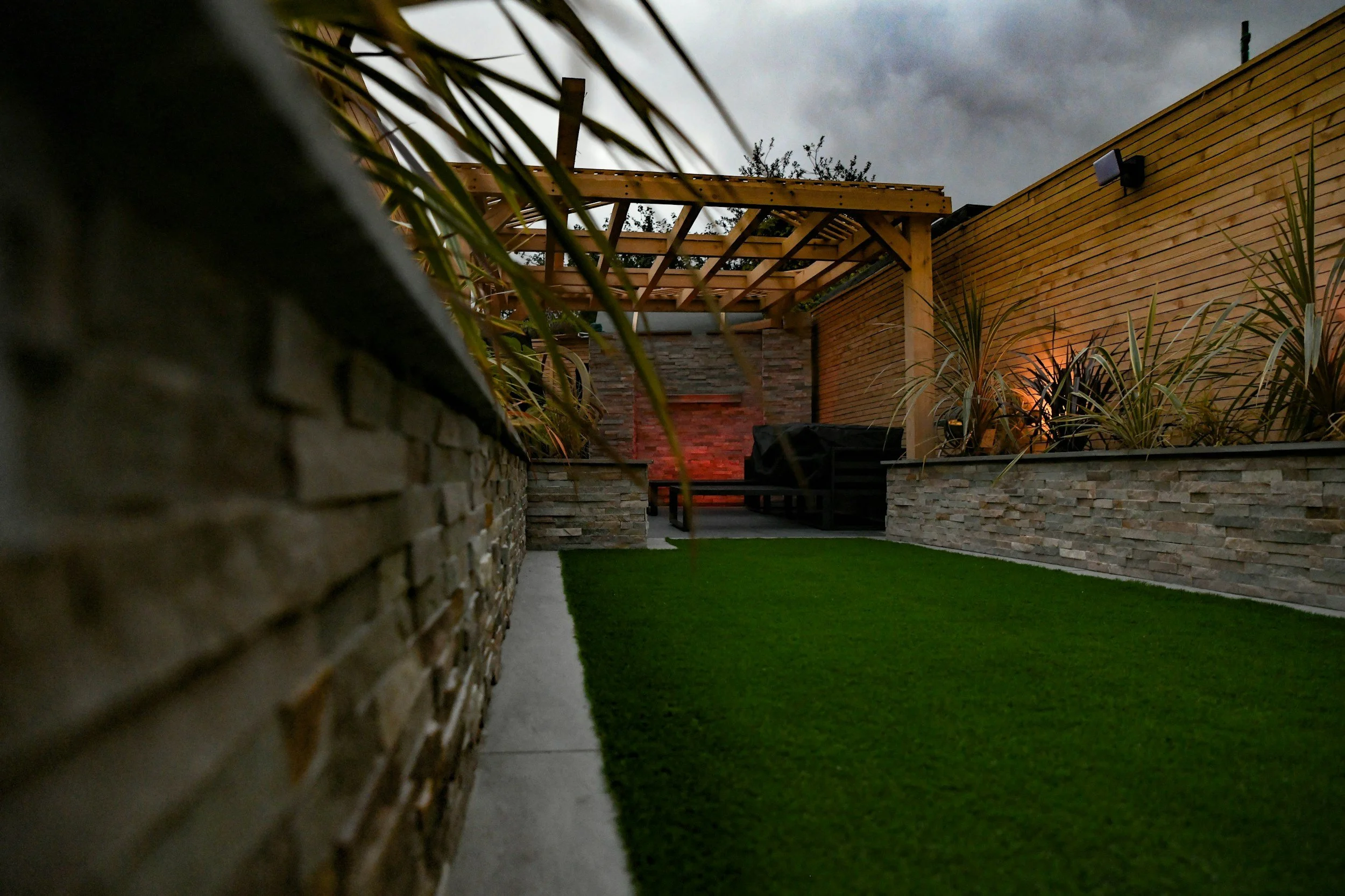 Nighttime view of a modern backyard with a stone wall, wooden pergola structure, and landscaped plants, with cloudy sky overhead.
