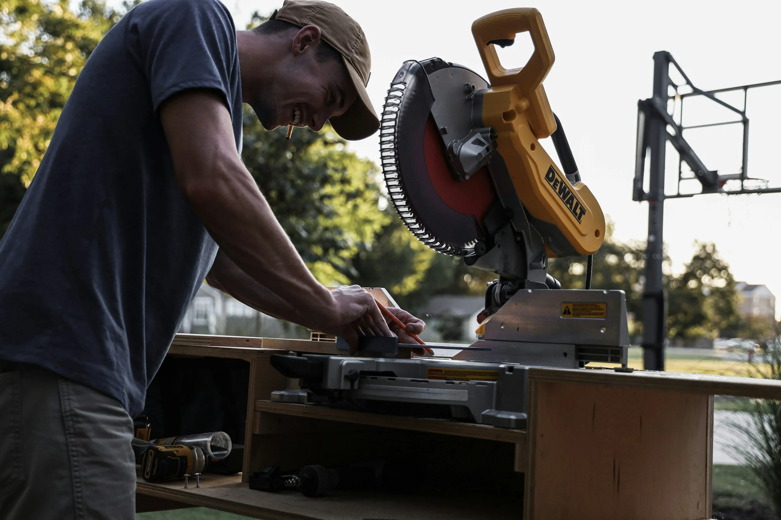 A man operating a DeWalt miter saw outdoors during sunset, cutting a piece of wood on a workbench.