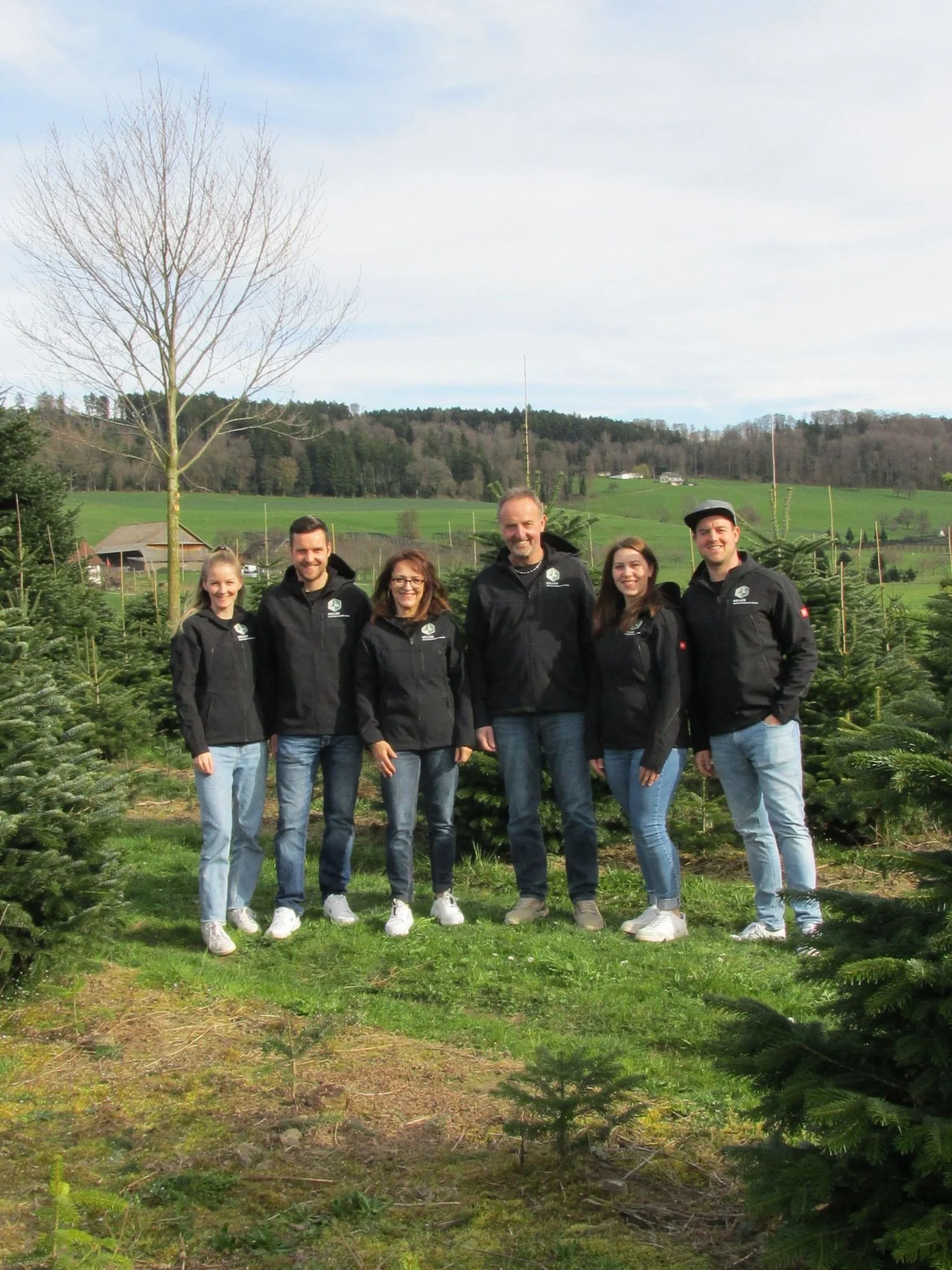 Eine Gruppe von sechs Menschen aus Keller Weihnachtsbäume GmbH stehen in einer Weihnachtsbaumplantage. Sie tragen schwarze Jacken und lächeln.