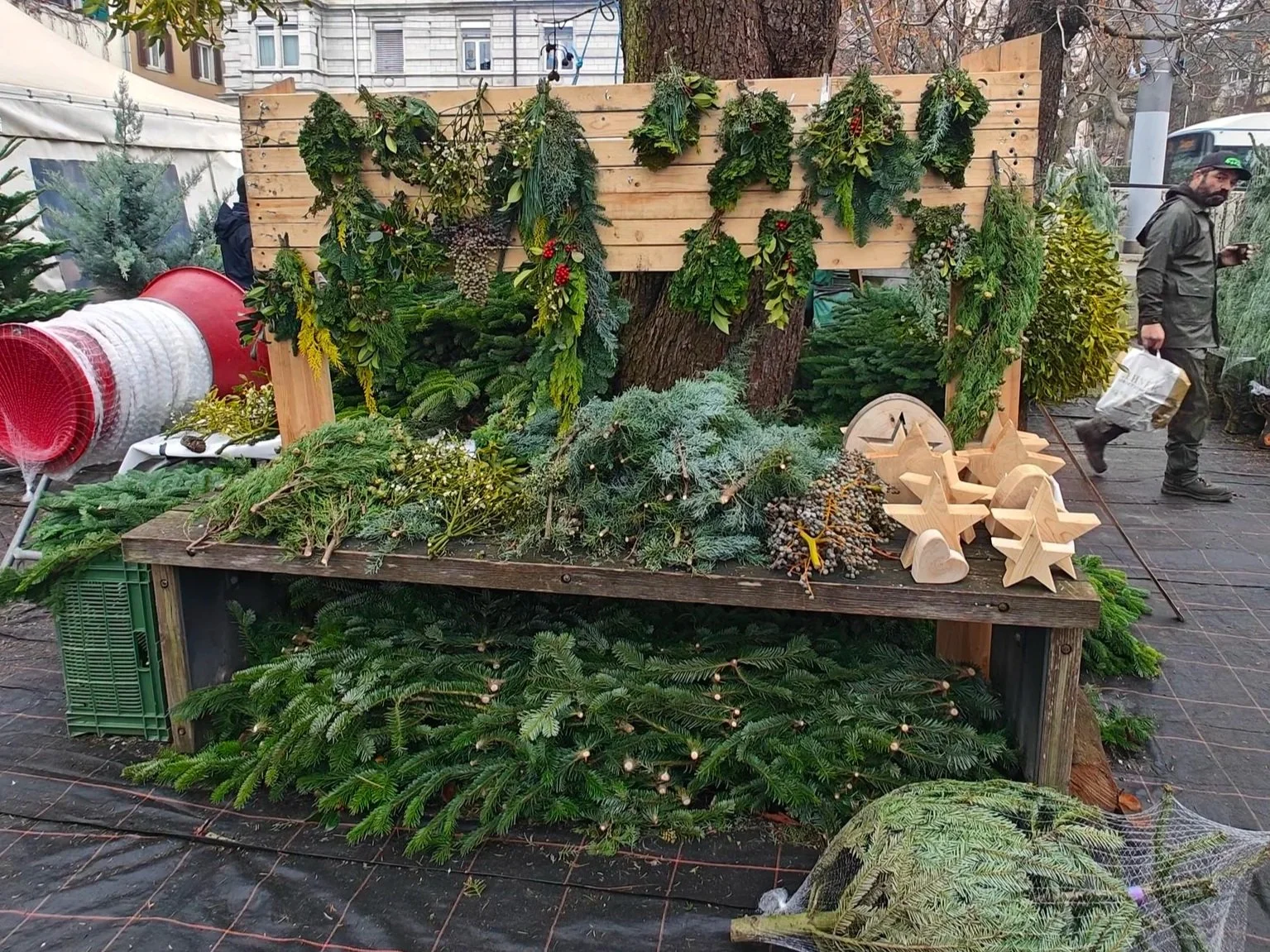 Ein Weihnachtsmarktstand mit Tannenzweigen, Kiefern- und Eukalyptussträußen, Holzdeko und Weihnachtskugeln, im Hintergrund sind Menschen und Gebäude sichtbar.