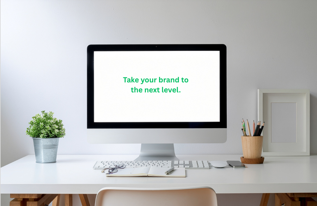 Computer monitor displaying the message 'Take your brand to the next level.' on a white background, surrounded by a potted plant, a notebook with glasses, a keyboard, and a container of pens, all on a white desk in a minimalist workspace.