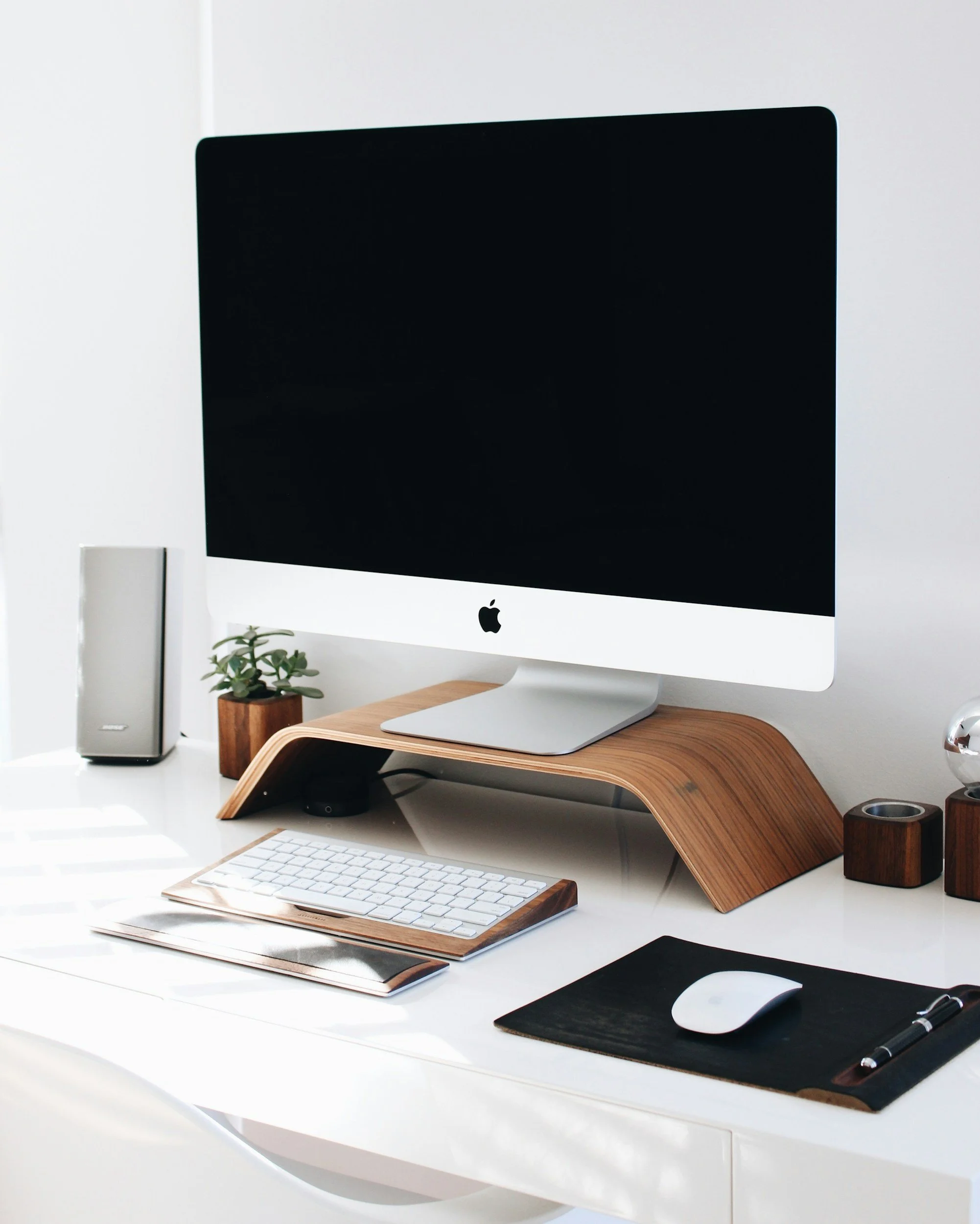 A modern workspace featuring an Apple iMac on a wooden stand, a wireless keyboard, a mouse on a black mouse pad, a small potted plant, a speaker, and a notebook with a pen, all on a white desk in a bright room.