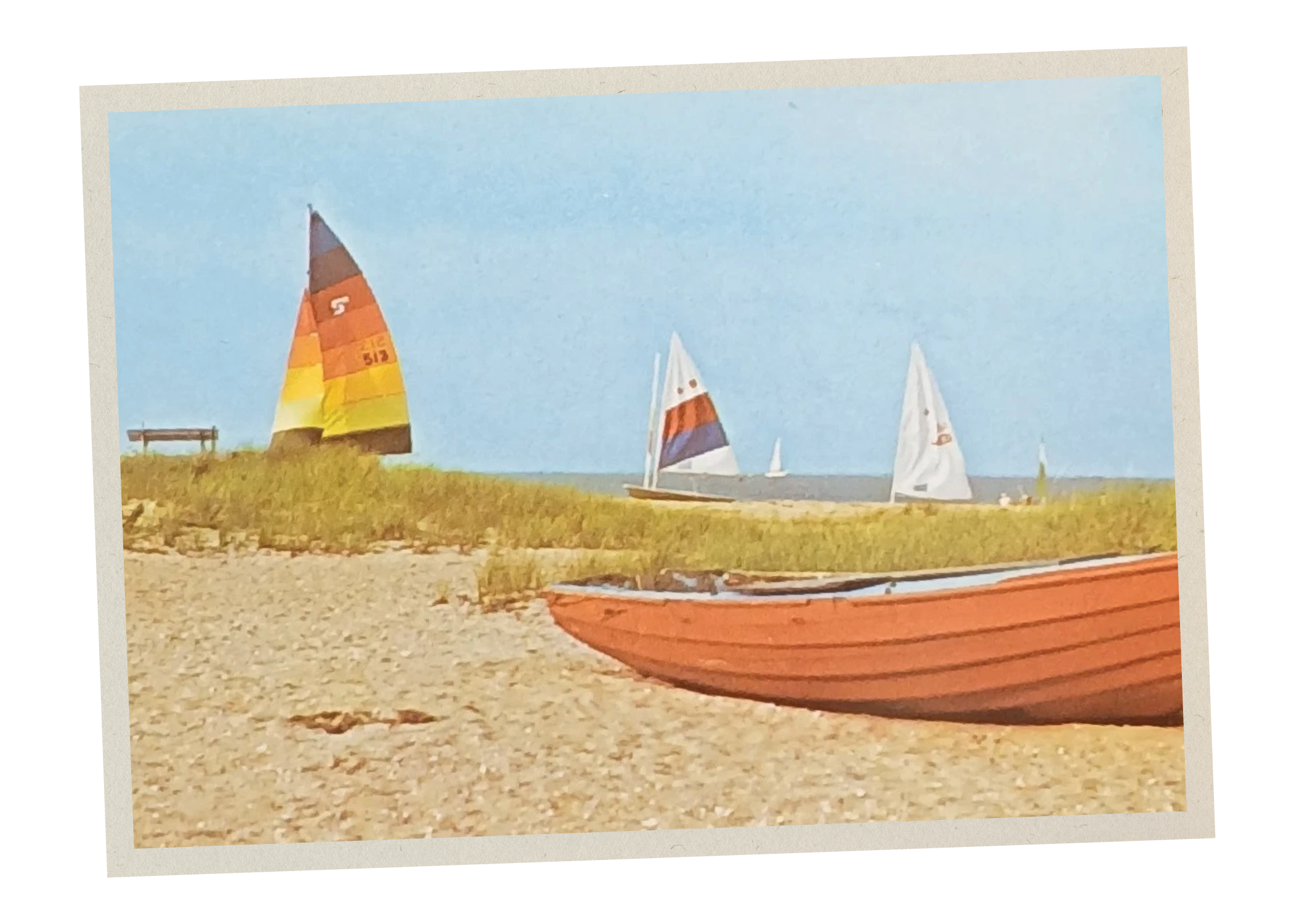 A red boat resting on a sandy beach with grass and sailboats in the background under a clear blue sky.