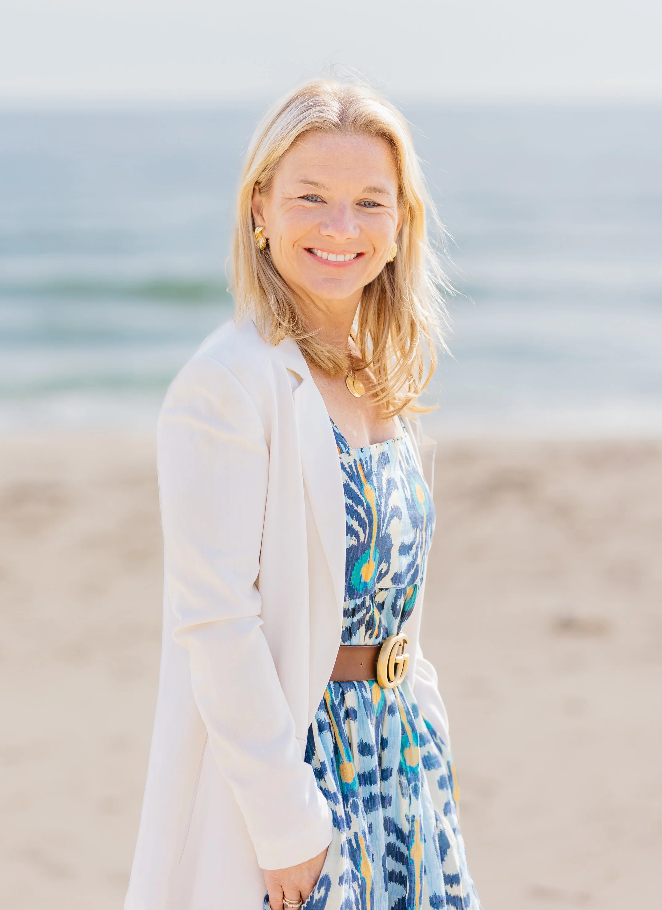 A smiling woman with blonde hair on a beach with the ocean in the background, wearing a white blazer, a colorful patterned dress, and gold jewelry.