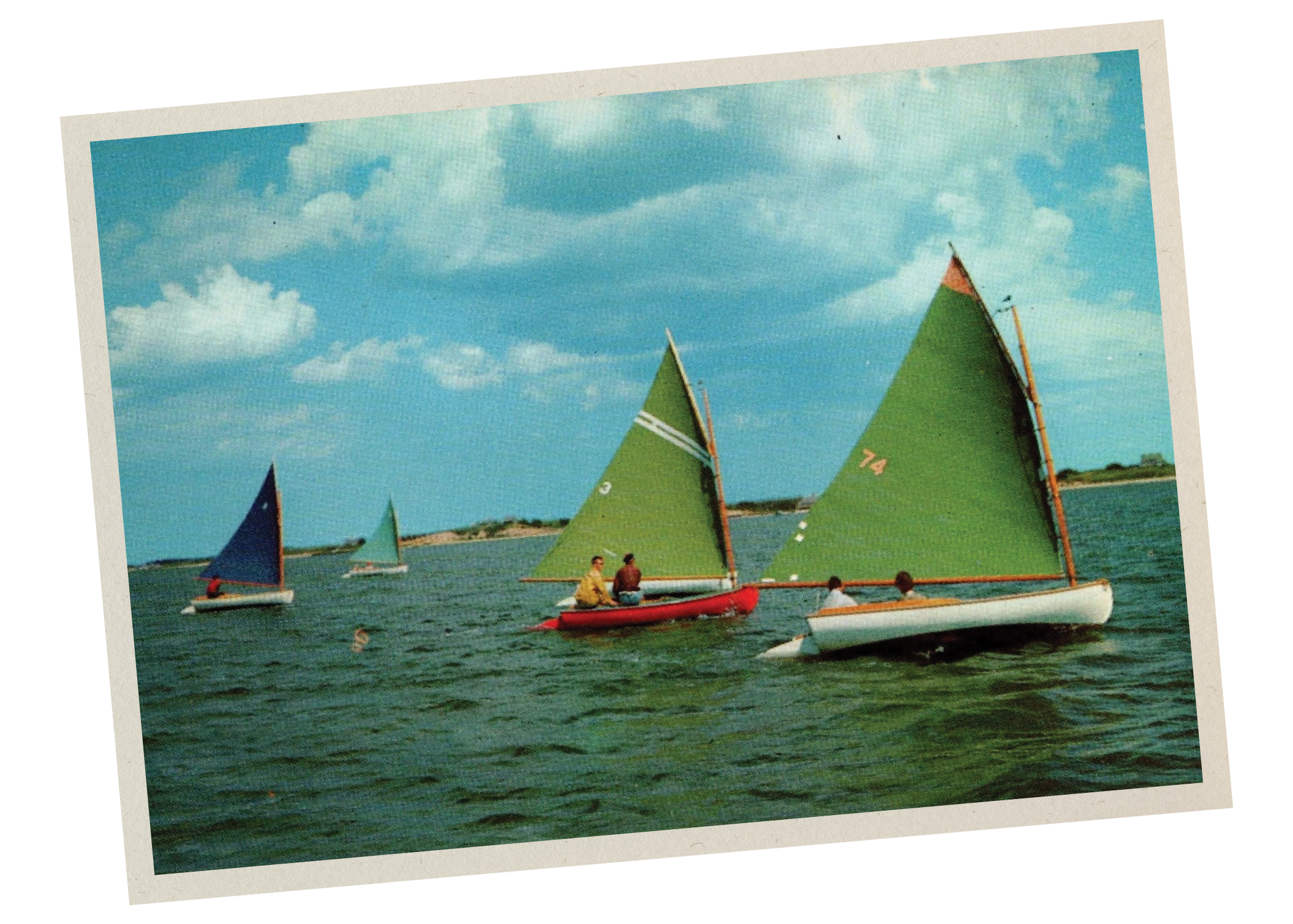 Color photograph of sailboats with colorful sails on the water, and two people sitting in a red boat in the foreground, with a cloudy sky in the background.