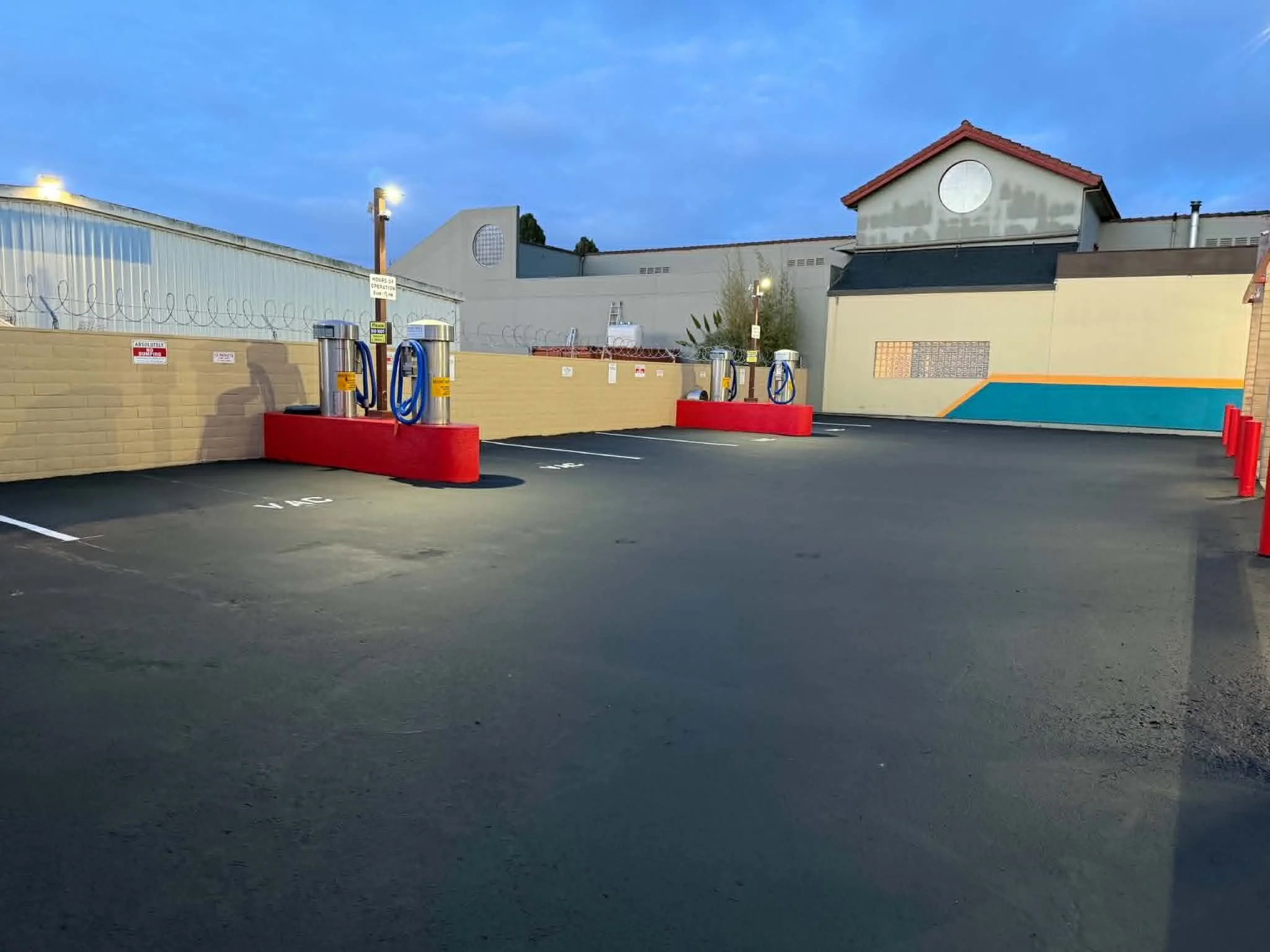 Empty parking lot with four electric vehicle charging stations, a brick wall, building with barbed wire, and a clear blue sky at dusk.