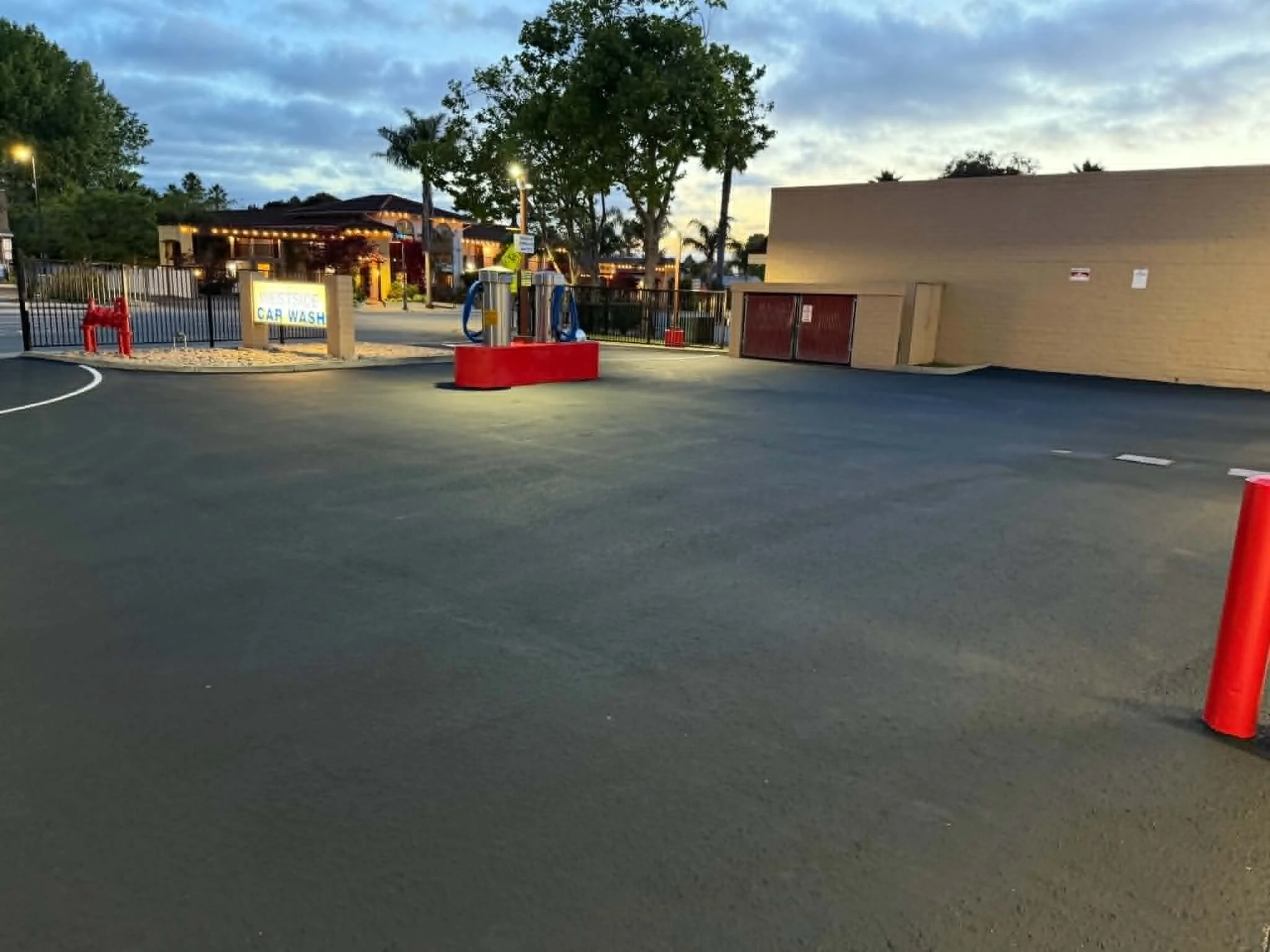 An outdoor car wash facility at dusk with a sign, soap dispensing equipment, trees, a building, and orange traffic cones.
