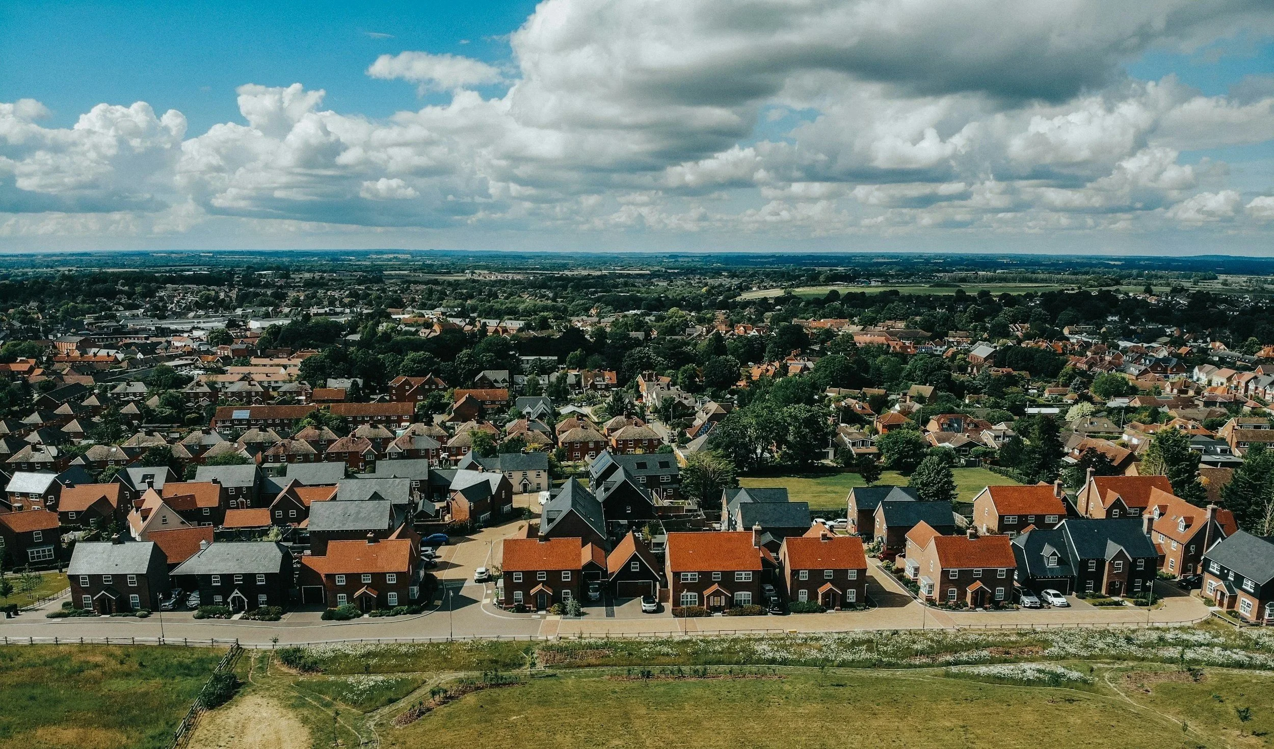 An aerial view of a suburban neighborhood with red brick houses, green trees, and streets under a partly cloudy sky.