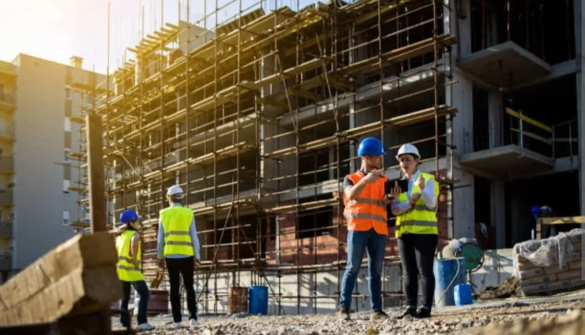 Construction workers inspecting plans on a construction site with a partially built multi-story building and scaffolding in the background.