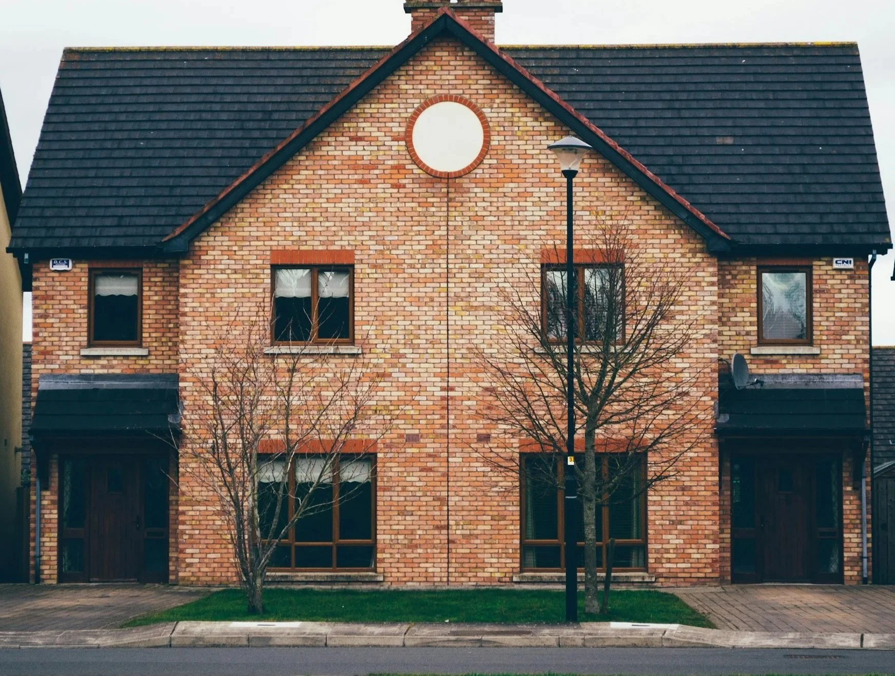 Front view of a brick house with a triangular roof, circular window on the upper gable, two rectangular windows with white curtains, and two main entrance doors, with a sidewalk, grass, trees without leaves, and a streetlamp in front.