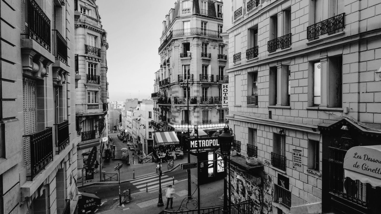 Black and white photo of a Parisian street corner with buildings, balconies, and signs for a hotel and cafes. Pedestrians and bicycles are visible on the street.