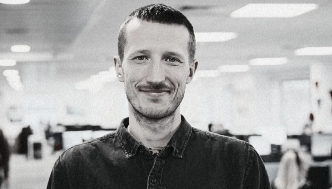 Black and white photo of a smiling man with short hair and a beard, wearing a button-up shirt, in an office setting.
