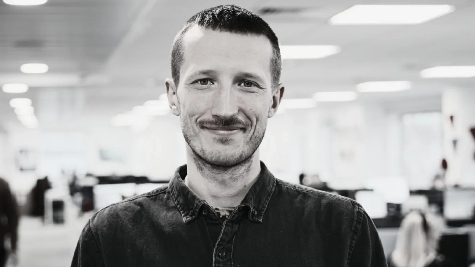 Black and white photo of a smiling man with short dark hair and a beard in an office environment.