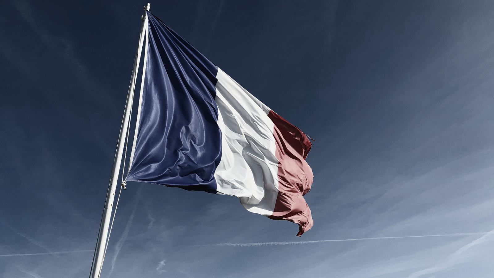 French flag waving in the wind against a blue sky with some clouds and contrails.