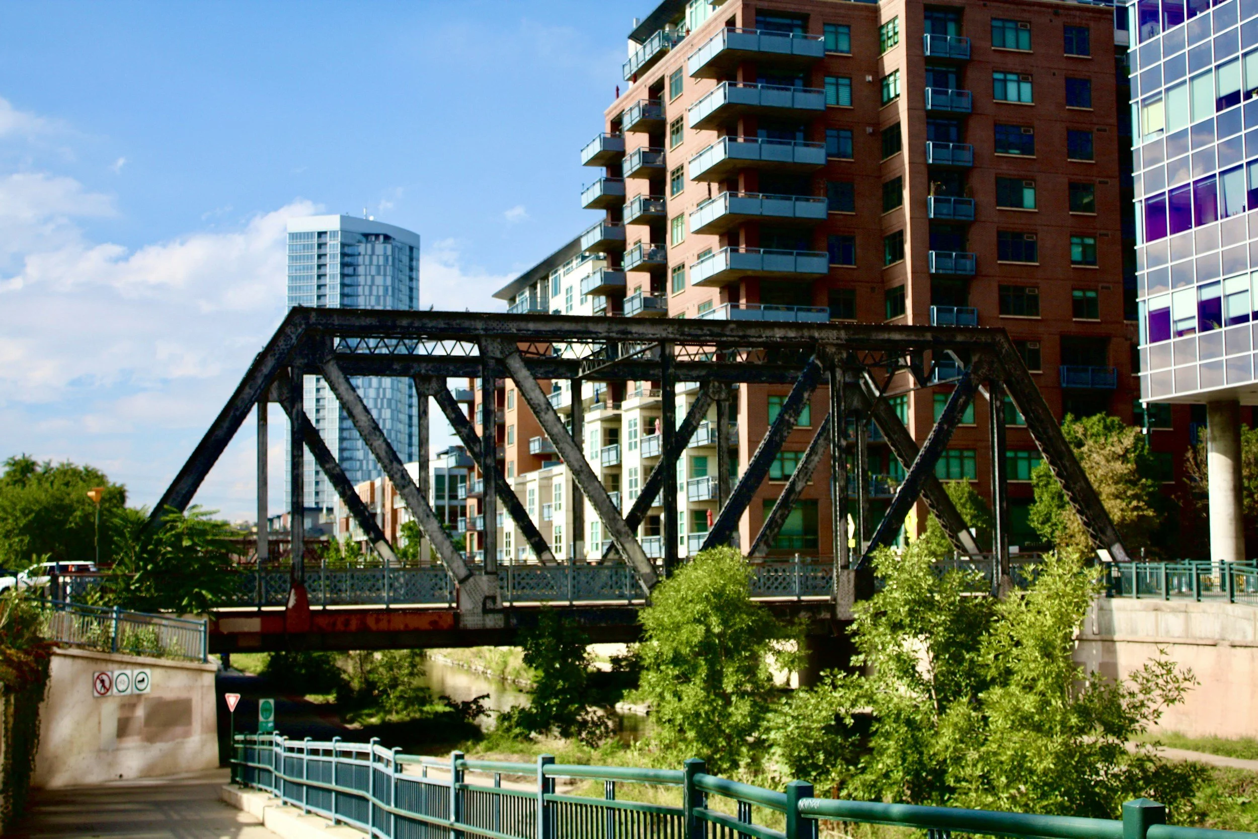 A cityscape with modern multi-story apartment buildings and a bridge over a river. The buildings have various colors and balconies. The sky is blue with scattered clouds.
