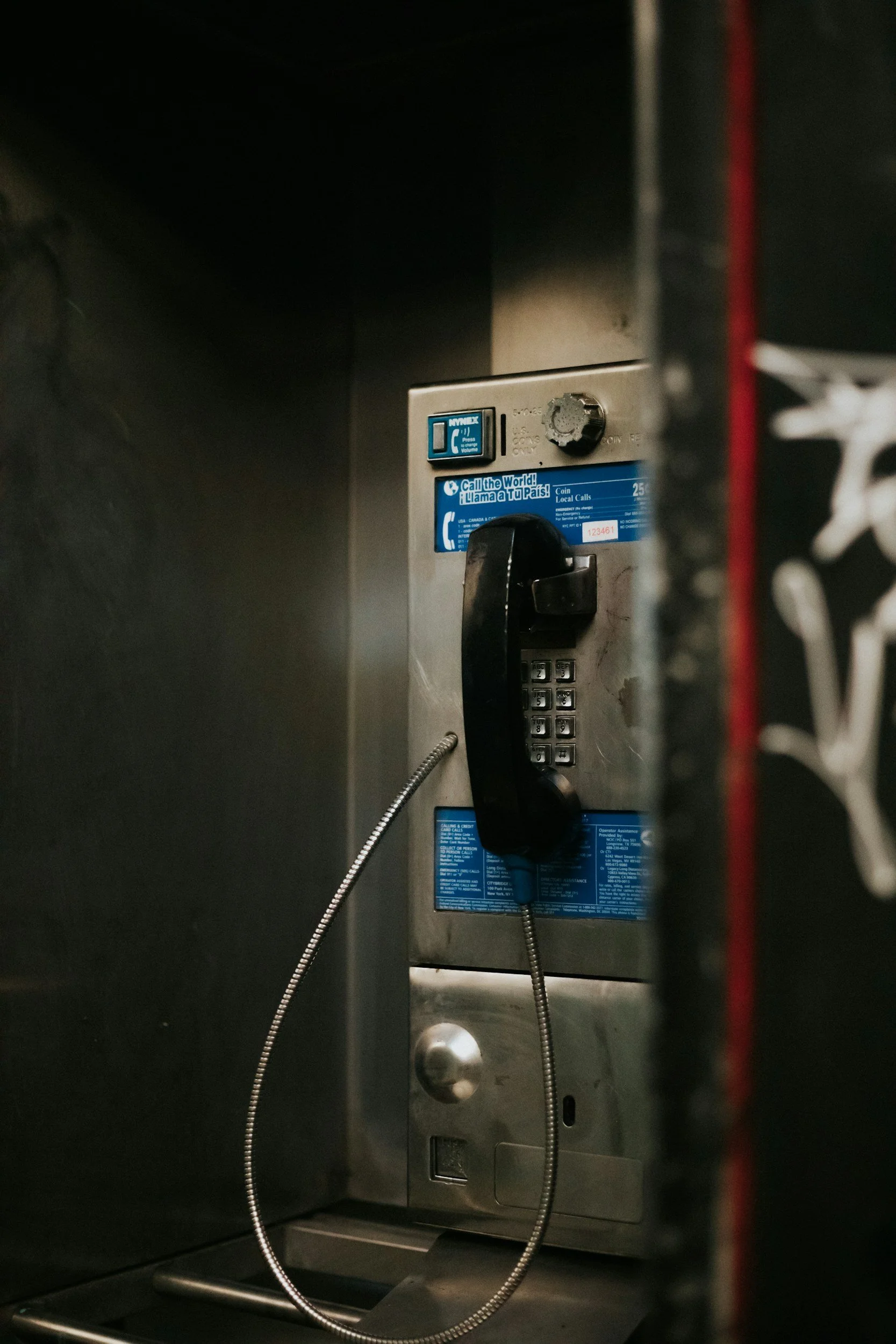 Inside a telephone booth with a black payphone, a coiled cord, and a blue informational sticker on the wall.