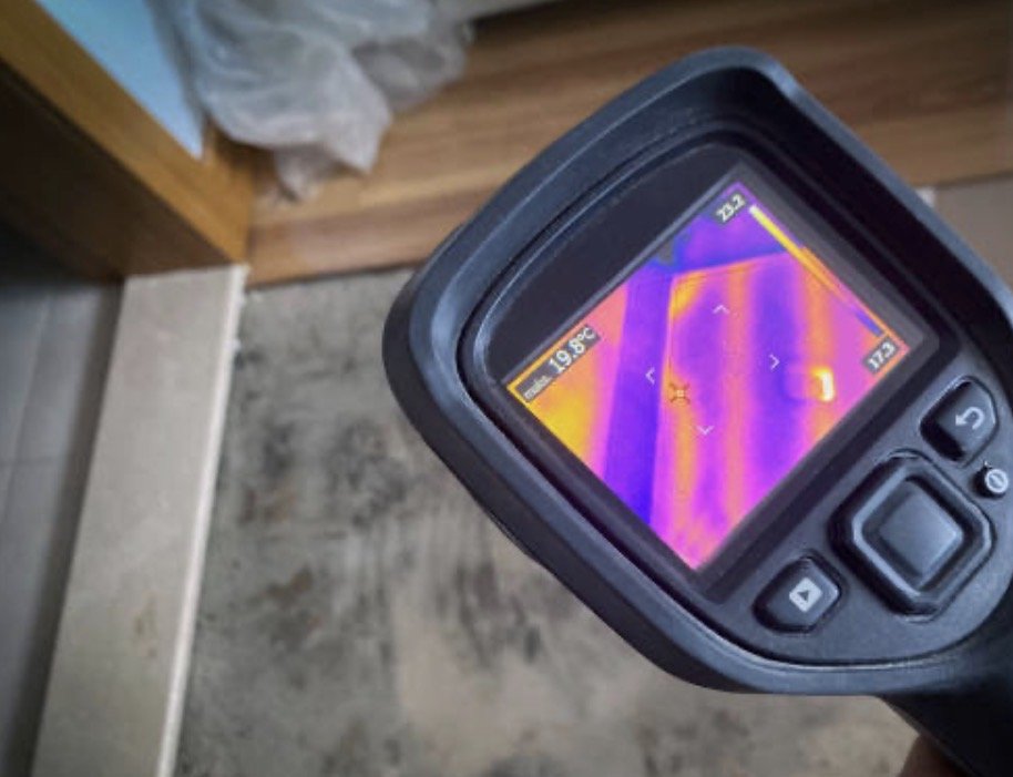 Thermal imaging camera showing heat patterns on a surface near a corner of a room, with the floor and wall in view.