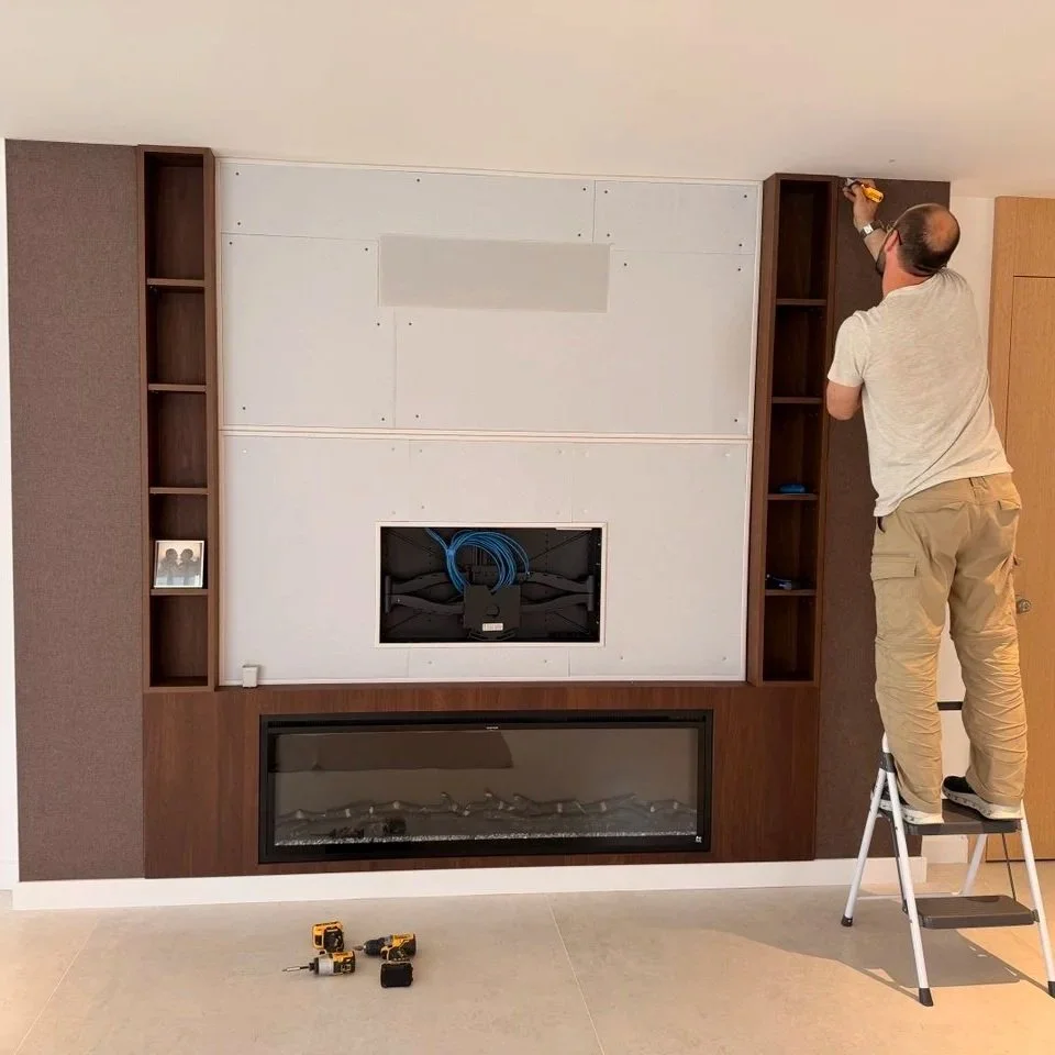A man standing on a step ladder installs or repairs a built-in wall unit with a fireplace below on a living room wall. The wall unit has open shelves on both sides and a large central space with cables inside.
