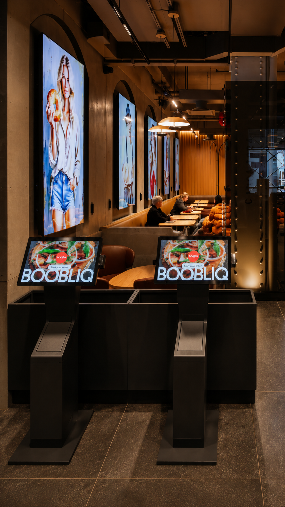Self-service ordering kiosks at a restaurant with digital screens displaying food images and the text "BOOBLIQ". In the background, a dining area with people sitting at tables, and large colorful posters of women on the wall.