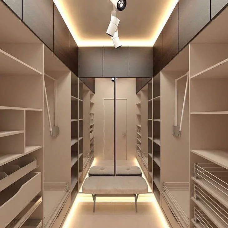 A walk-in closet with built-in white shelving on both sides, dark cabinets at the top, a mirror on the back wall, and a beige bench in the center. Recessed lighting illuminates the space.