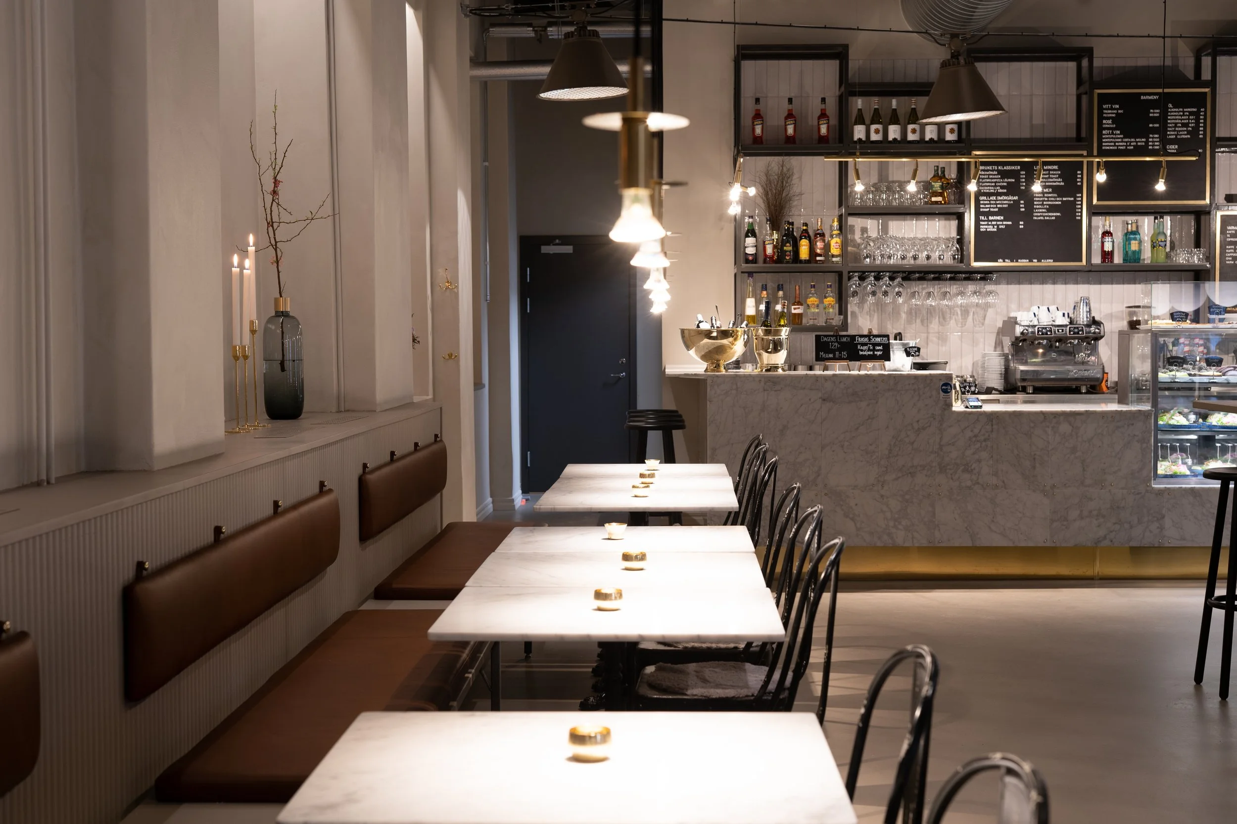 En modern restaurang eller café med vita marmorbord, svarta stolar och varm belysning. På vänster sida finns tallrikar och ett vackert arrangemang av ljus och grenar i en vas. Bakom disken finns ett sortiment av alkohol, glas och maskiner för kaffe och läsk.
