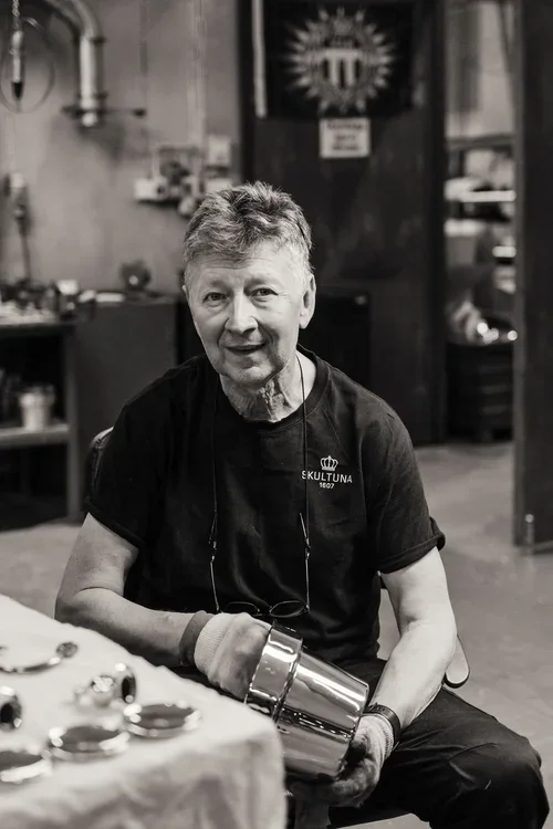 En äldre man med grått hår och svart T-shirt som sitter vid ett bord i ett rum, håller en metallkanna, i bakgrunden syns dekorativa föremål och en vägg med en symbol.