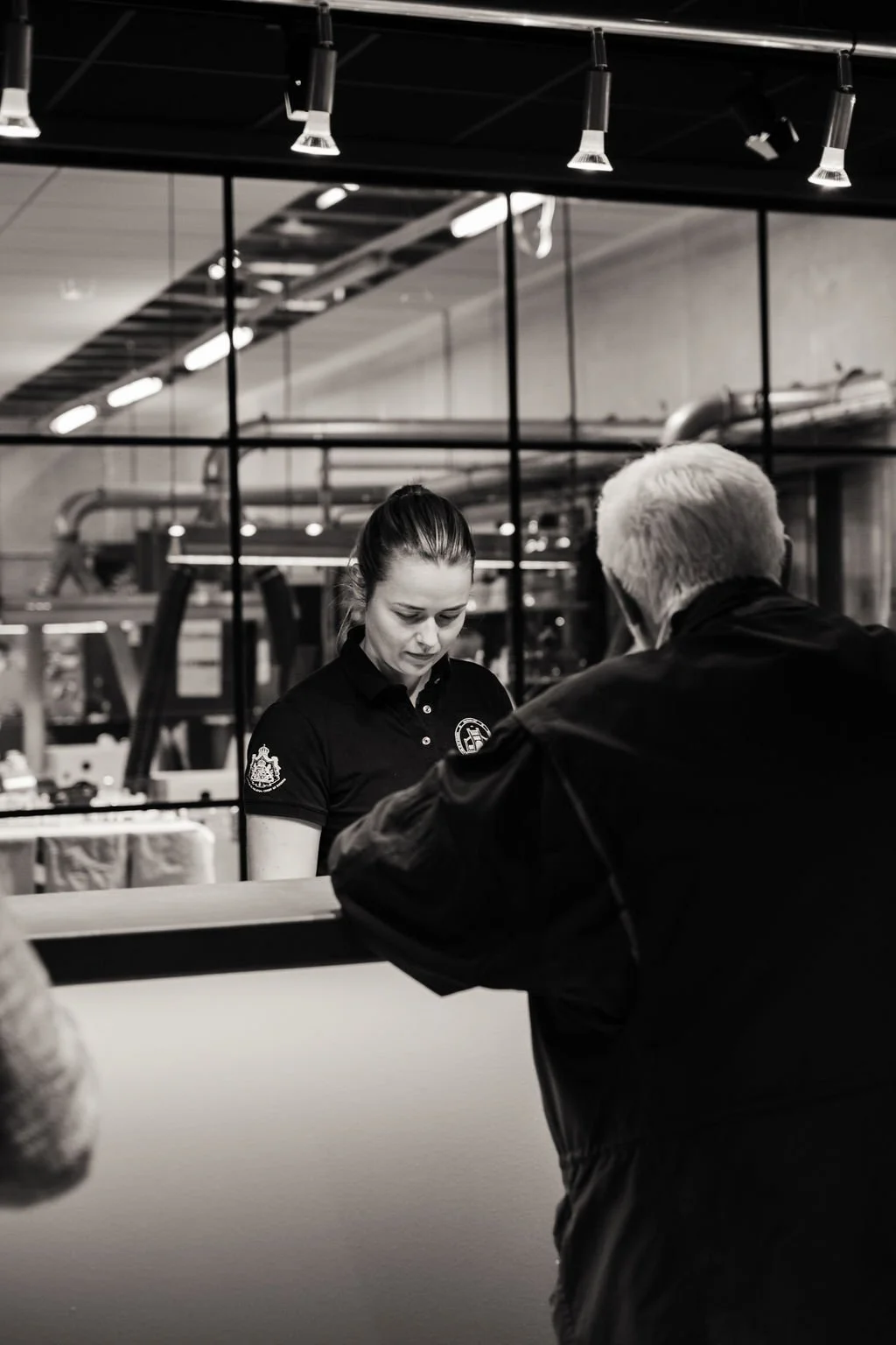 En svartvit bild av en ung kvinna bakom en bardisk som pratar med en äldre man, i ett industriellt inrett kafé eller bar.