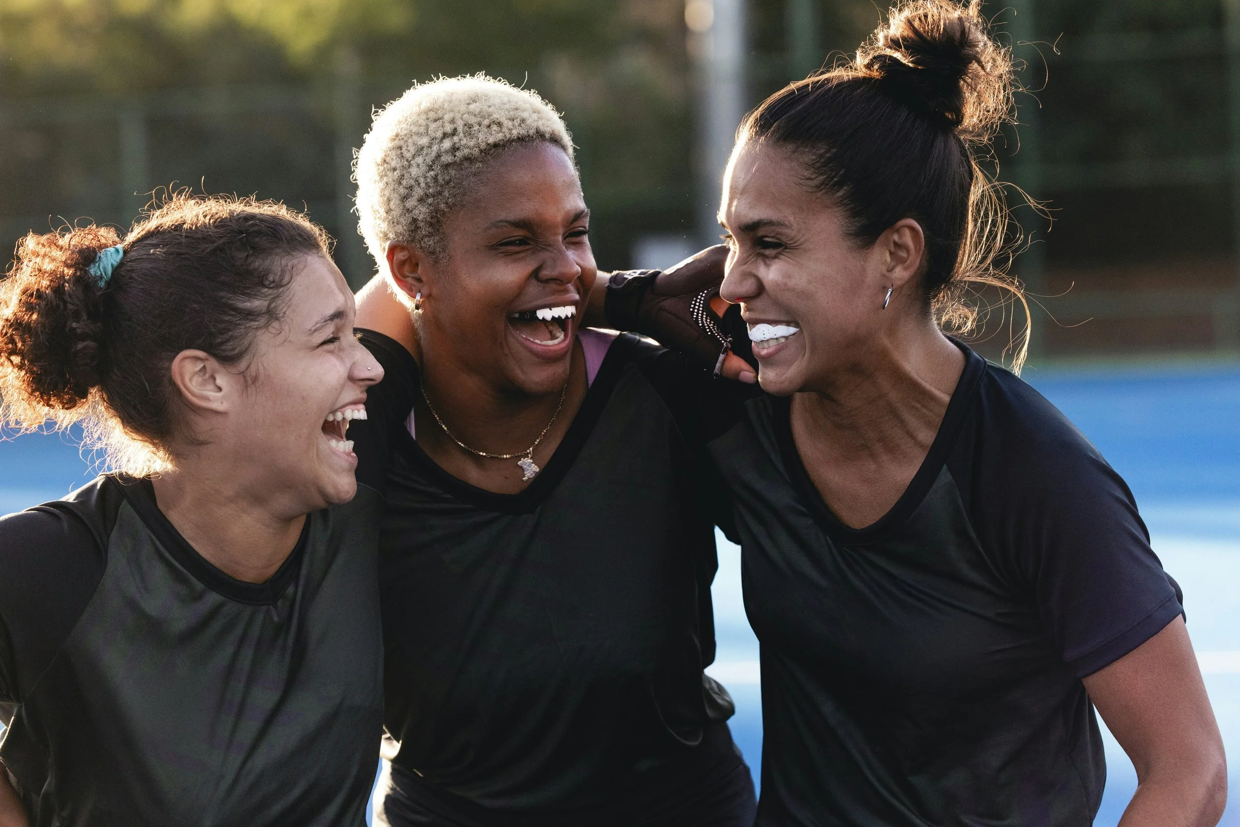 Three people in athletic clothing are standing on a tennis court, smiling and talking to each other. They are outdoors with trees and a fence visible in the background.