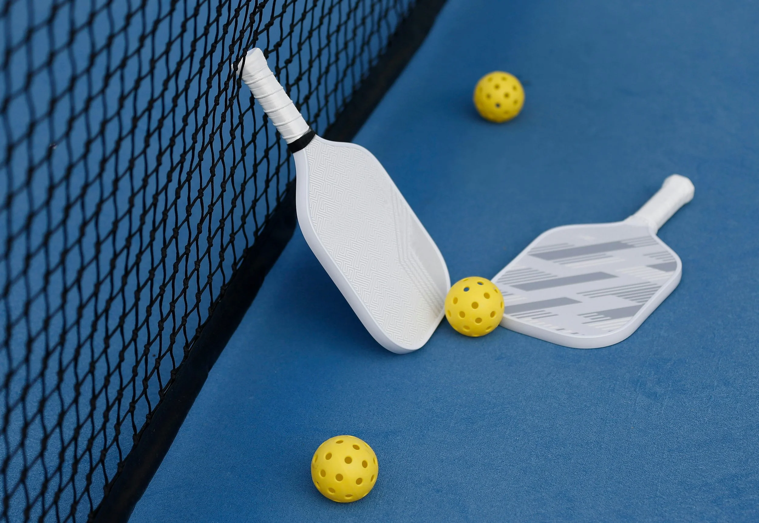 A tennis court with a black mesh net, two yellow perforated balls, a white tennis racket, and a white plastic chair on a blue surface.