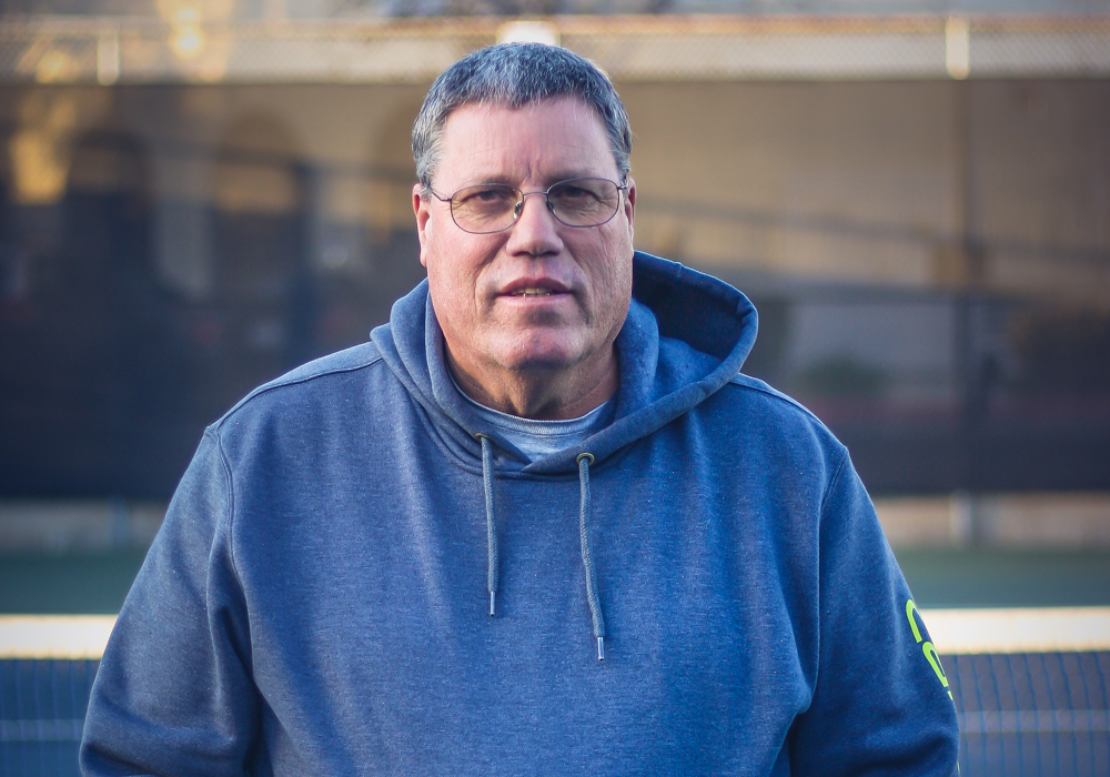 A middle-aged man wearing glasses and a blue hoodie standing on a tennis court.