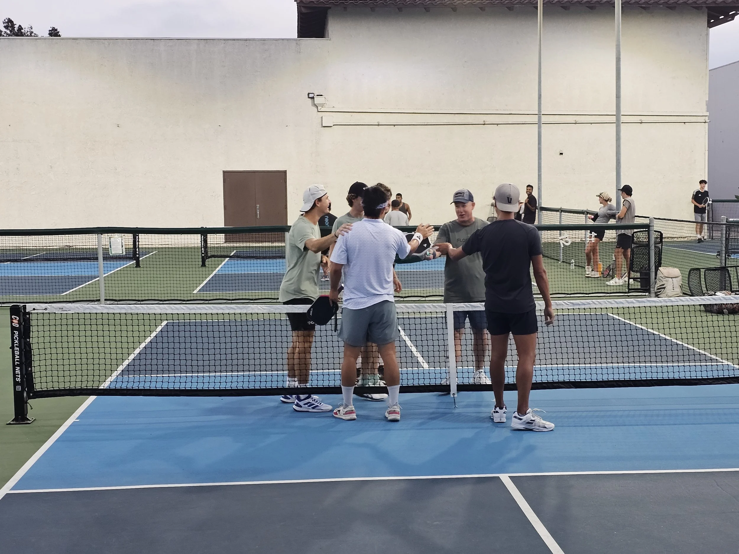 A group of young people playing tennis on a court, some chatting near the net, with others in the background, overcast sky, large beige building in the background.