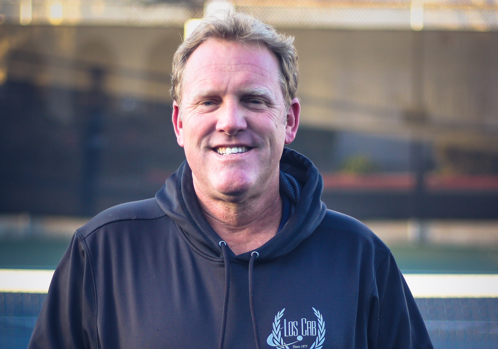 A smiling man wearing a black hoodie with a Los Cab logo, standing outdoors near a sports field.