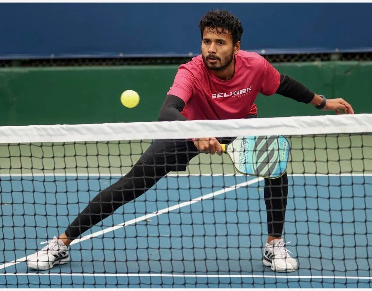A man playing pickleball on an indoor court, wearing a red shirt and black leggings, preparing to hit a yellow ball with a paddle.