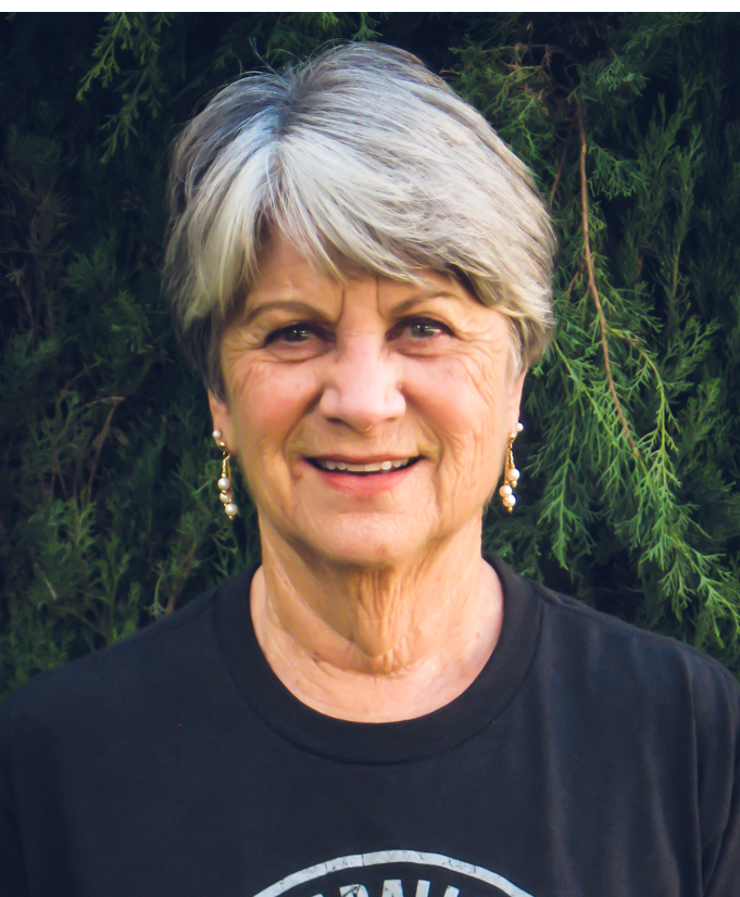 A smiling elderly woman with short gray hair and earrings, standing in front of green foliage.