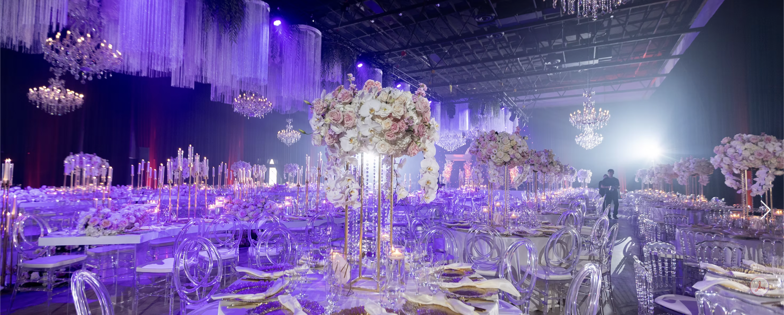 Elegant event space decorated with tall floral centerpieces and numerous candles, with purple lighting and chandeliers hanging from the ceiling.