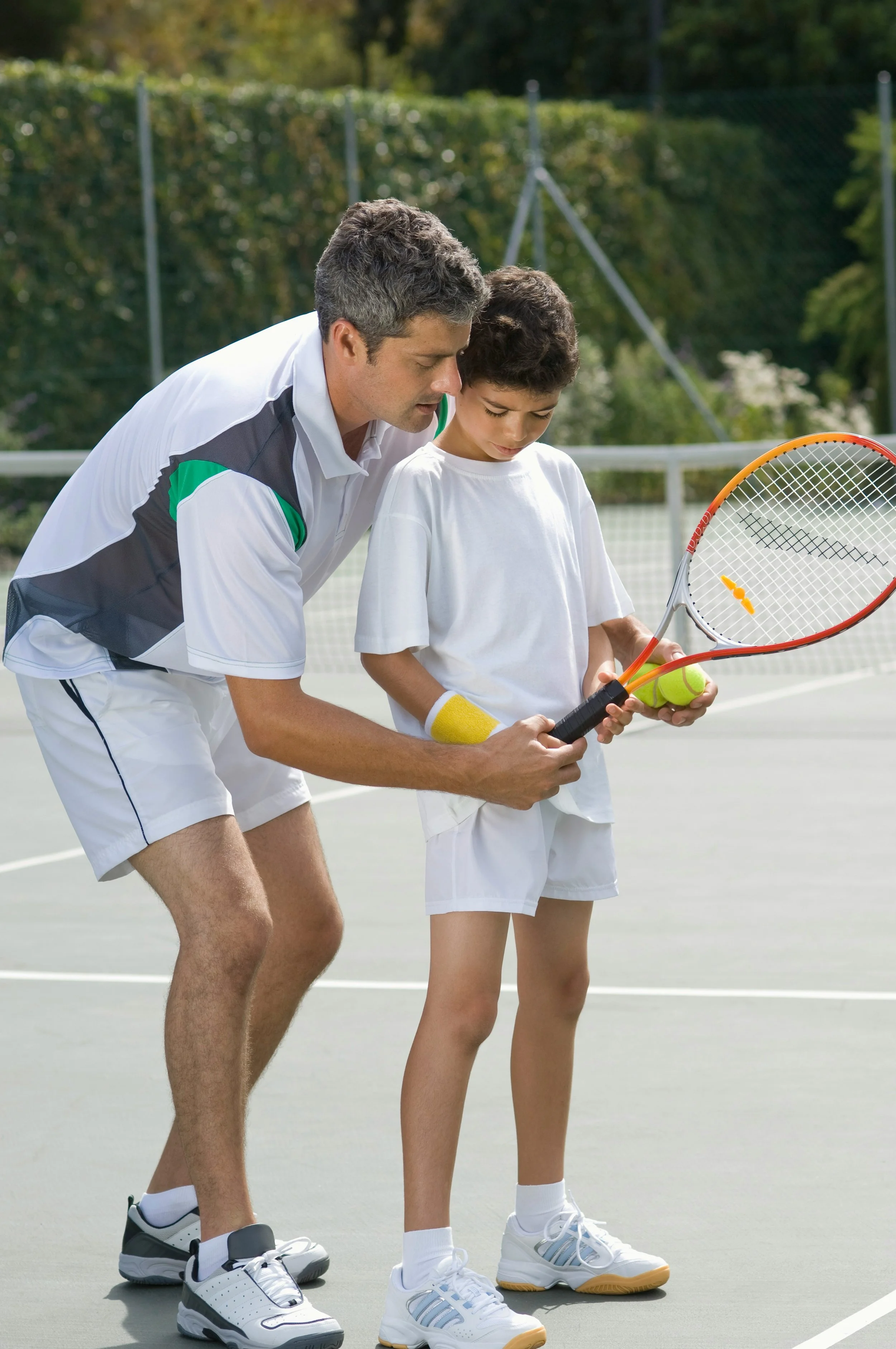 A coach helping a young boy with tennis on an outdoor tennis court, guiding him on how to hold a tennis racket, with tennis balls in his hand, surrounded by a green background and fence.