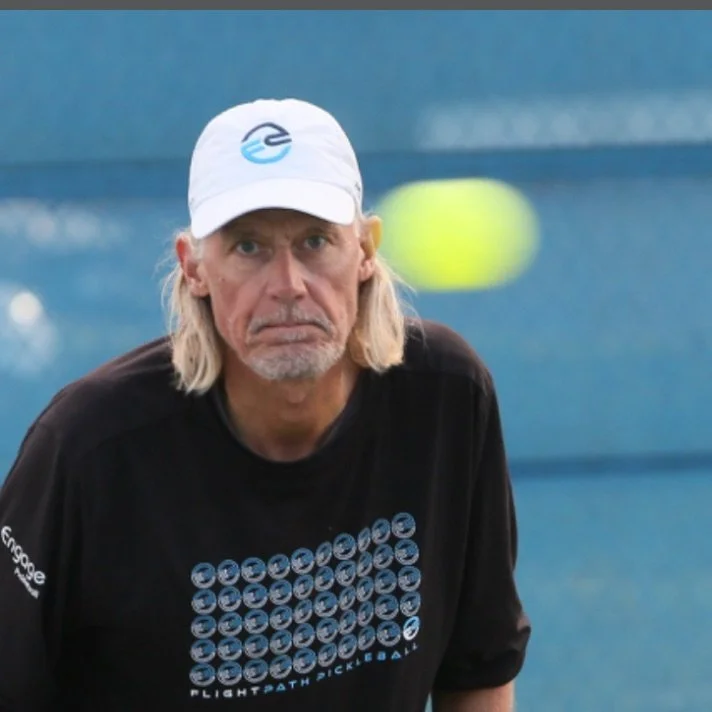 An older man with long, gray hair and a beard, wearing a white cap and black shirt, standing on a tennis court with a blurred tennis ball in the background.