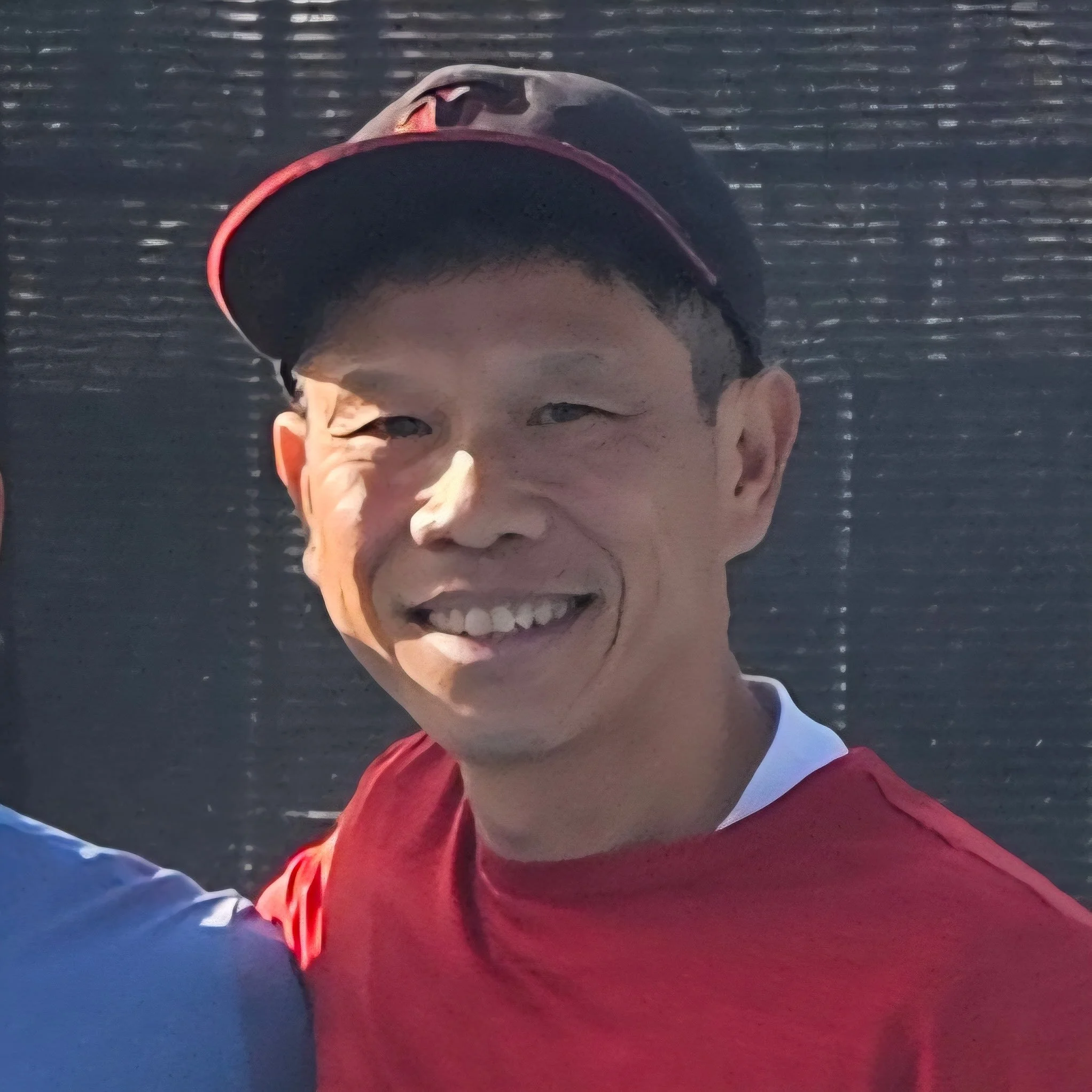 A young man wearing a red and blue jacket and a black and red cap, smiling, with a dark background and slight reflections.