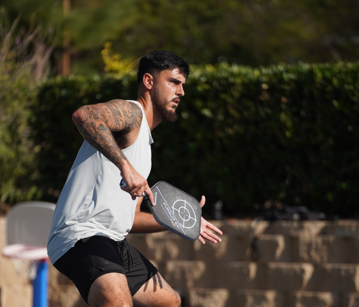 Man with tattoos playing pickleball outdoors on a sunny day, holding a paddle and preparing to hit the ball.