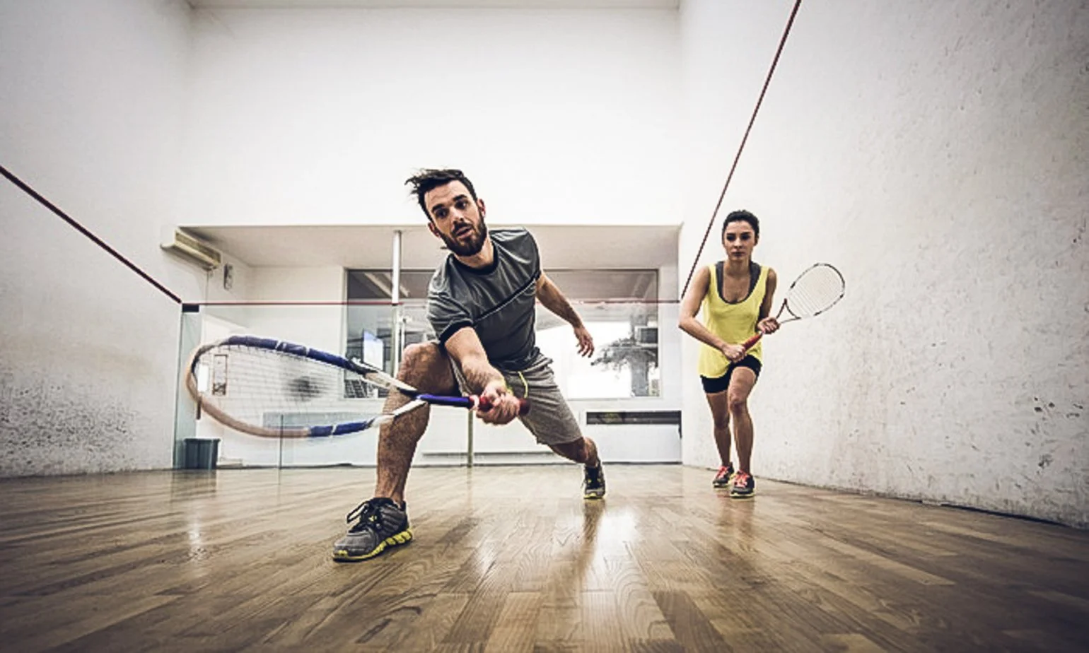 A man and a woman playing squash on an indoor court, both holding squash rackets, with the man lunging forward and the woman running behind him.