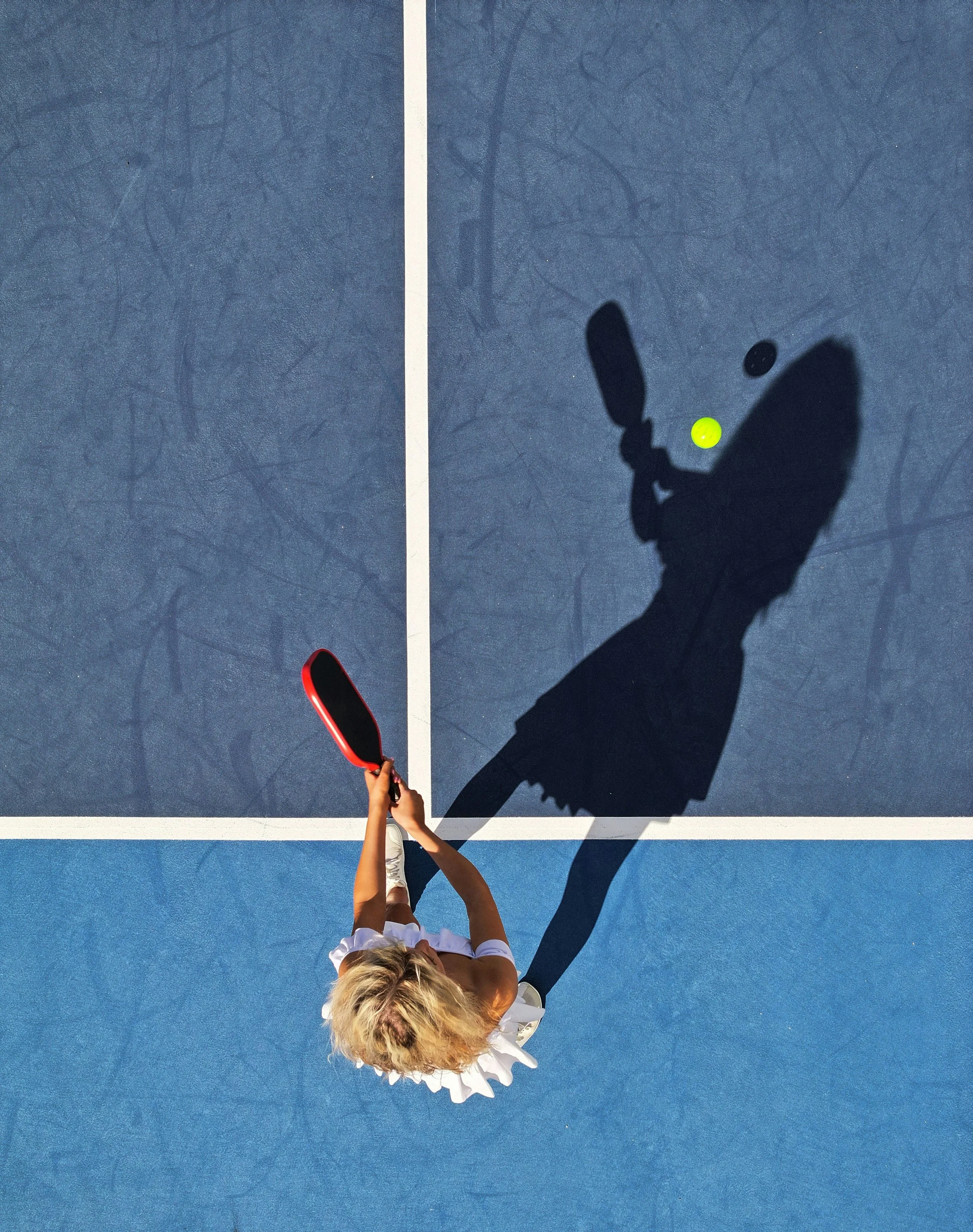 Top-down view of a young woman with blonde hair playing tennis on a blue tennis court. She is wearing a white shirt and a white ruffled skirt, holding a red tennis racket, with a green tennis ball near her racket. The court has a white baseline and sidelines, and her shadow is cast on the court.