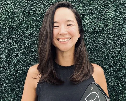 A young woman with shoulder-length dark hair smiling in front of a green leafy background.